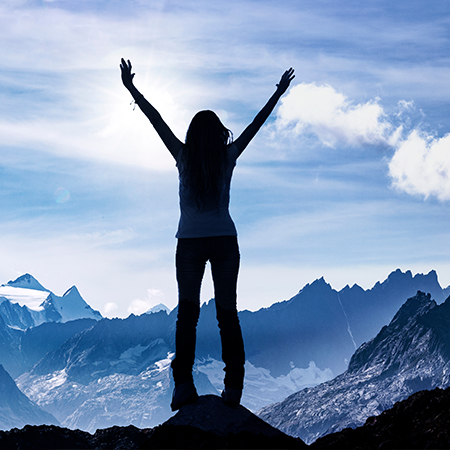 Die Silhouette einer Frau, die auf einem Berg steht, beide Arme in die Höhe streckt und in die Ferne auf einen strahlend blauen Himmel und weitere Bergkämme blickt